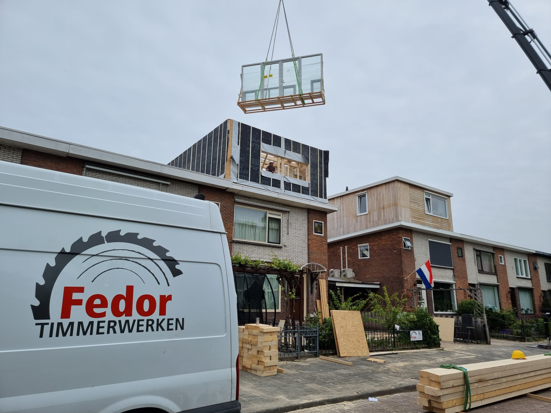 Bus van Fedor Timmerwerken terwijl een kraan kozijnen naar een renovatieproject hijst